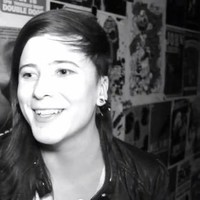 A black and white photo of Cam at an art show; it is a candid photo of them smiling during an interview. They are a white person with freckles and long straight hair that is cropped short on one side.
